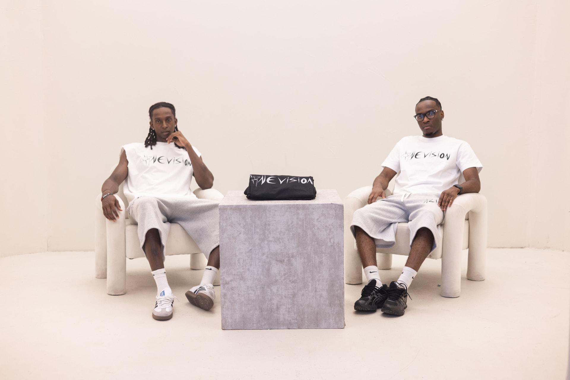 Two models in white 1VISION tees seated in bone armchairs either side of a concrete plinth